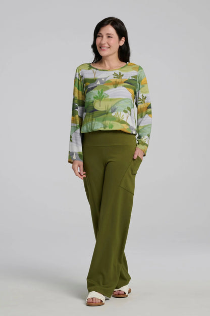A woman wearing the Belica Blouse by Kollontai in a Granny Smith toned nature print. It has a wide neck, long sleeves, linen-look fabric, and an elasticized waist for a subtle blouson effect She is wearing it with the Susan Pants in Granny Smith and standing in front of a grey background. 