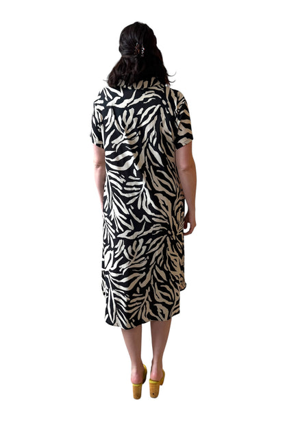 Back view of a woman wearing the Regina Dress by Compli K in Black and White print, a long dress with a v placket neck, short sleeves, loose fit, rounded hem and side slits. She is standing against a white background. 
