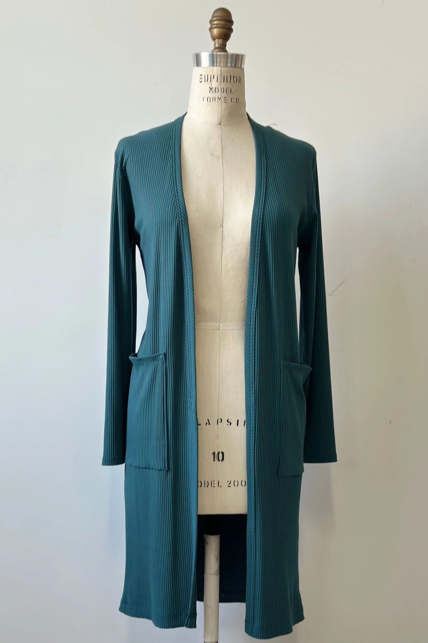 The Streliski Cardigan from Kollontai in Teal, a long open cardigan in bamboo rib with large patch pockets, is shown on a mannequin in front of a beige background. 