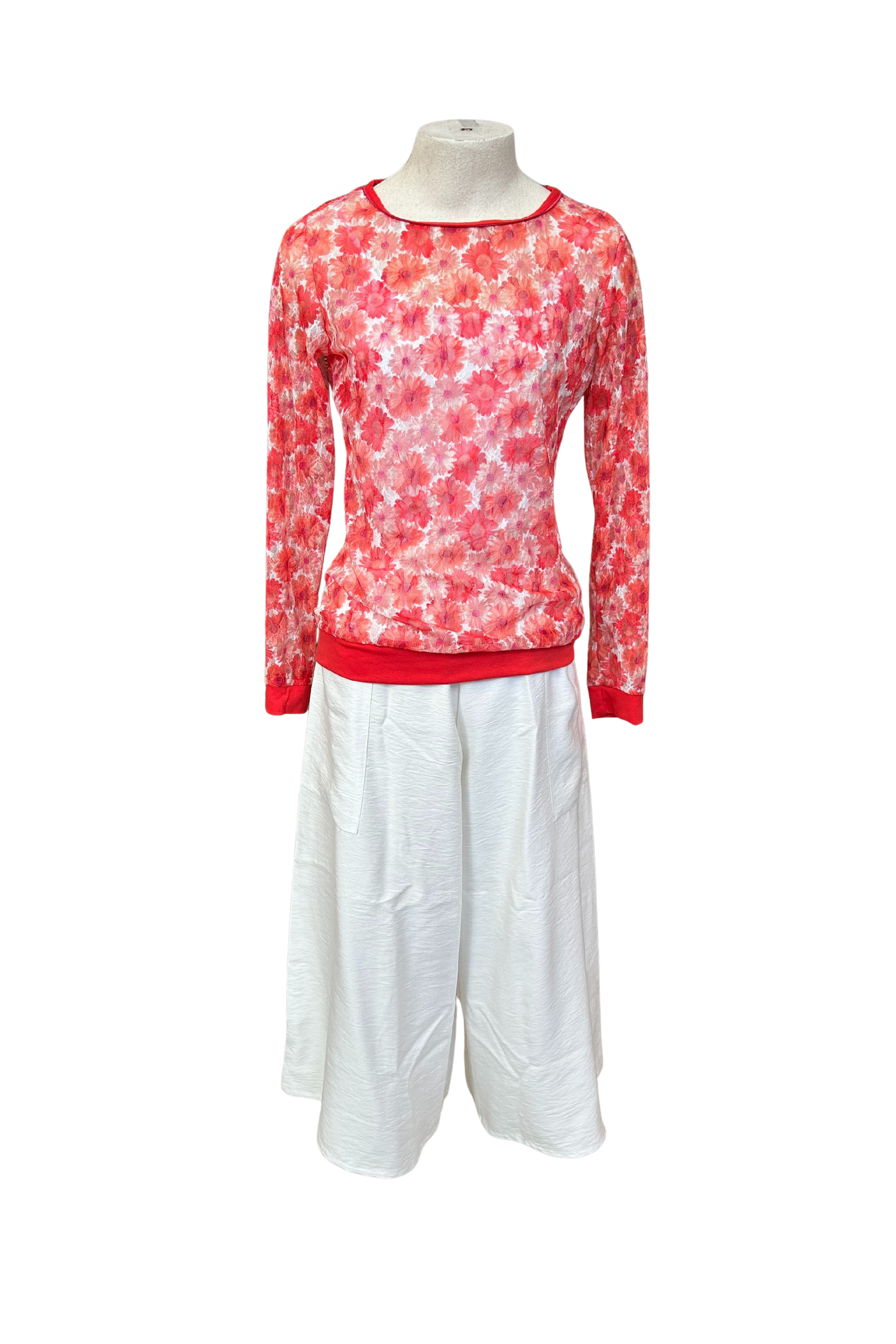 The Daisy Lace Shirt by Karkass in Coral, a floral lace shirt with solid cuffs and hem, is shown over a white tank and with white pants against a white background. 