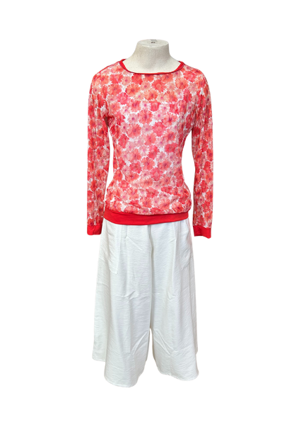 The Daisy Lace Shirt by Karkass in Coral, a floral lace shirt with solid cuffs and hem, is shown over a white tank and with white pants against a white background. 