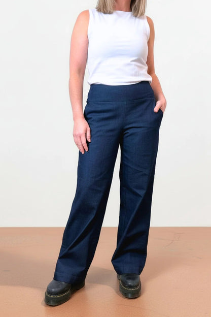 Neck down view of a woman wearing the Catherine Denim Pants by Message Factory, with a smooth pull-on waistband, wide legs, and pockets. She is wearing them with a white top and standing in front of a plain background.