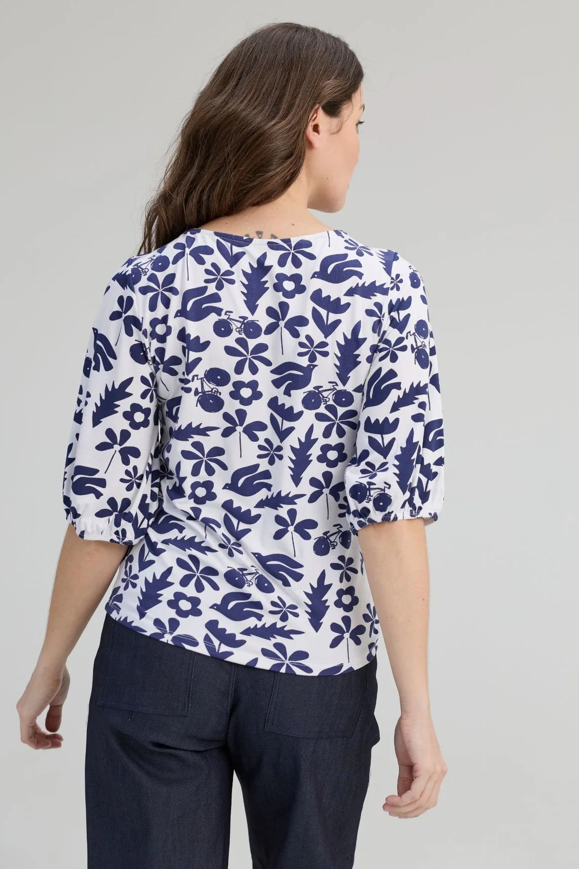 Back view of a woman wearing the Cadieux Top from message Factory in a floral Navy Print, with 3/4 sleeves with gathered cuffs, a wide neck and slim fit.  She is wearing it with dark pants and standing against a plain background. 