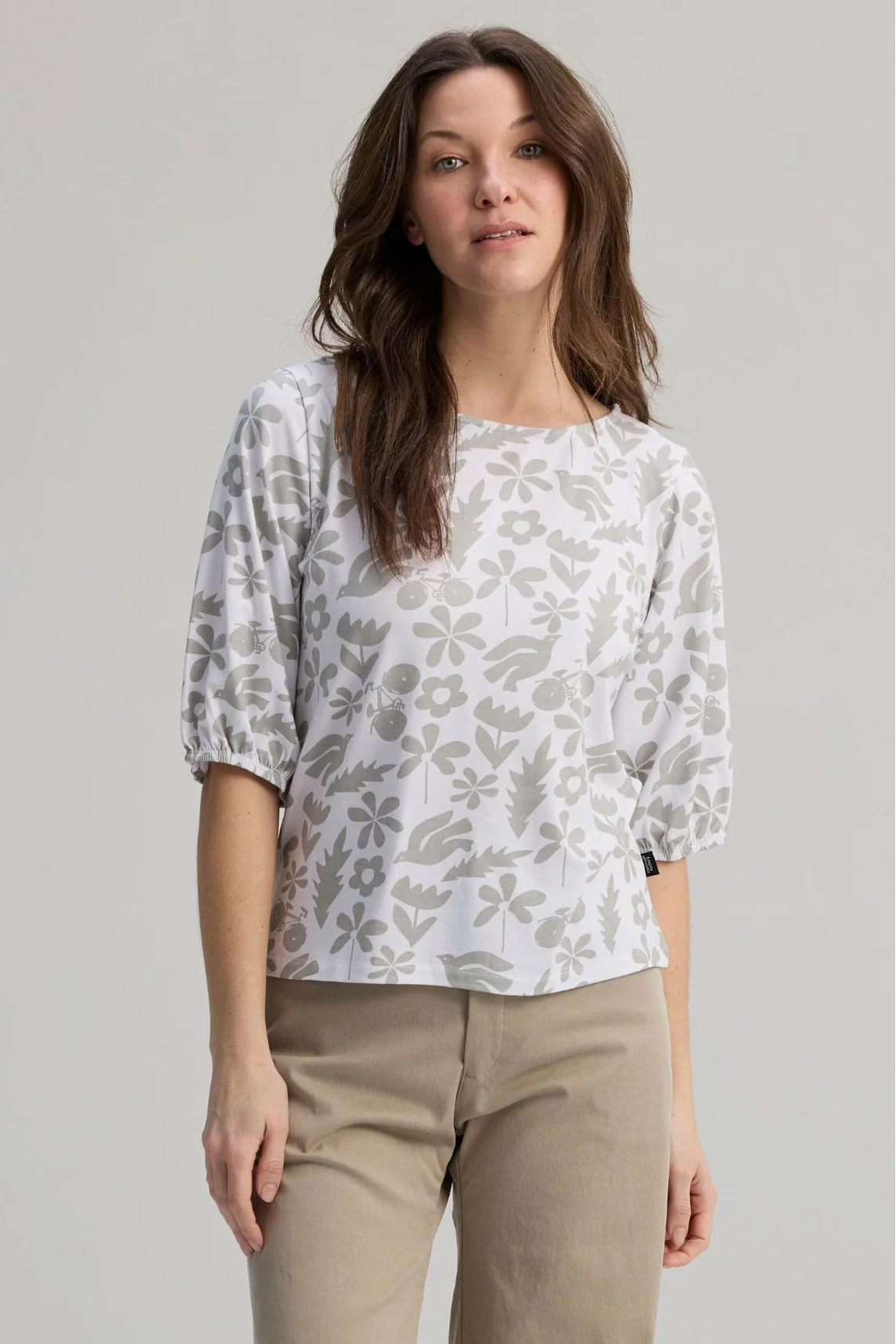 A woman wearing the Cadieux Top from Message Factory in a floral White Print, with 3/4 sleeves with gathered cuffs, a wide neck and slim fit.  She is wearing it with tan pants and standing against a plain background. 