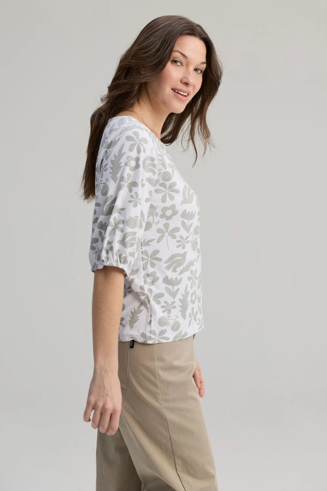 Side view of a woman wearing the Cadieux Top from Message Factory in a floral White Print, with 3/4 sleeves with gathered cuffs, a wide neck and slim fit.  She is wearing it with tan pants and standing against a plain background. 