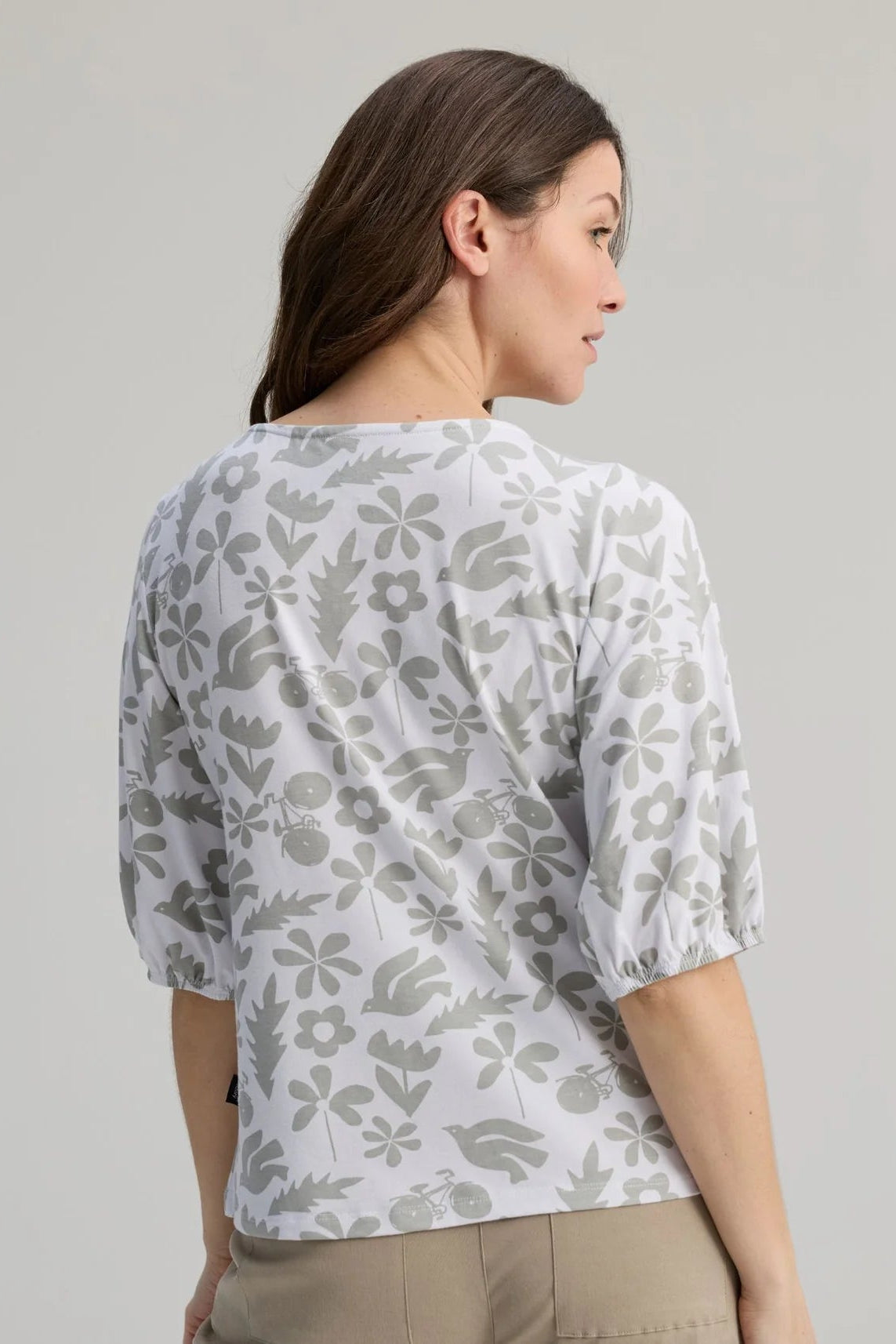Back view of a woman wearing the Cadieux Top from Message Factory in a floral White Print, with 3/4 sleeves with gathered cuffs, a wide neck and slim fit.  She is wearing it with tan pants and standing against a plain background. 