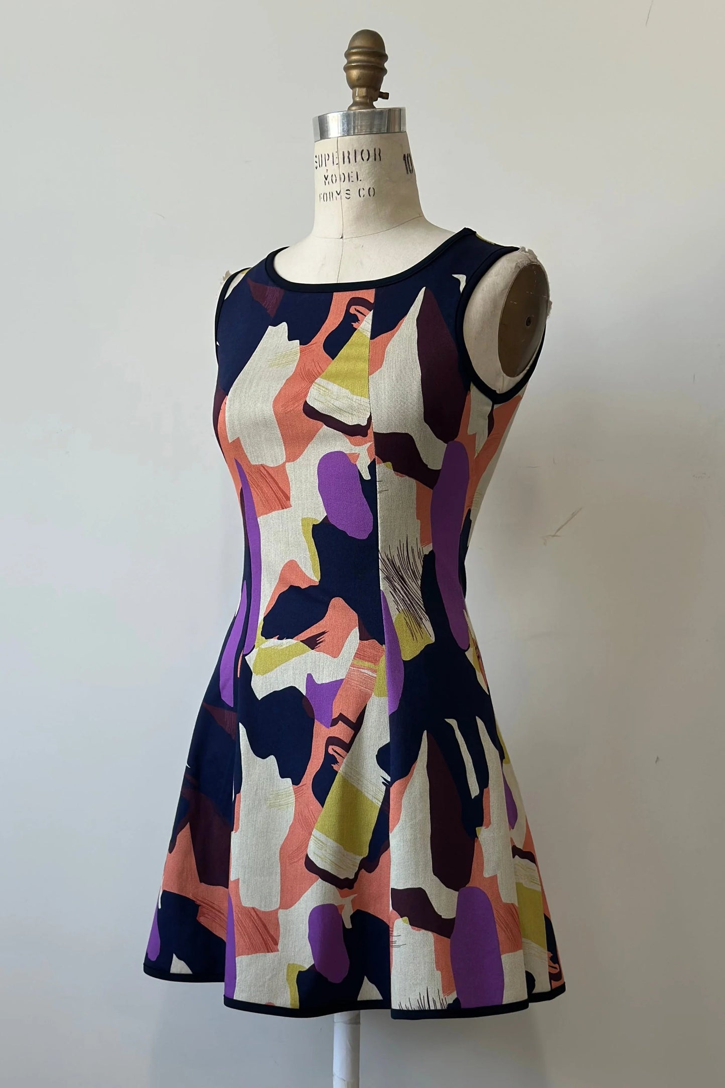 The Silba 2026 Dress by Kollontai, a sleeveless short dress in a Multicolour abstract print with a fit and flare shape and princess seams, is shown on a mannequin against a white background. 