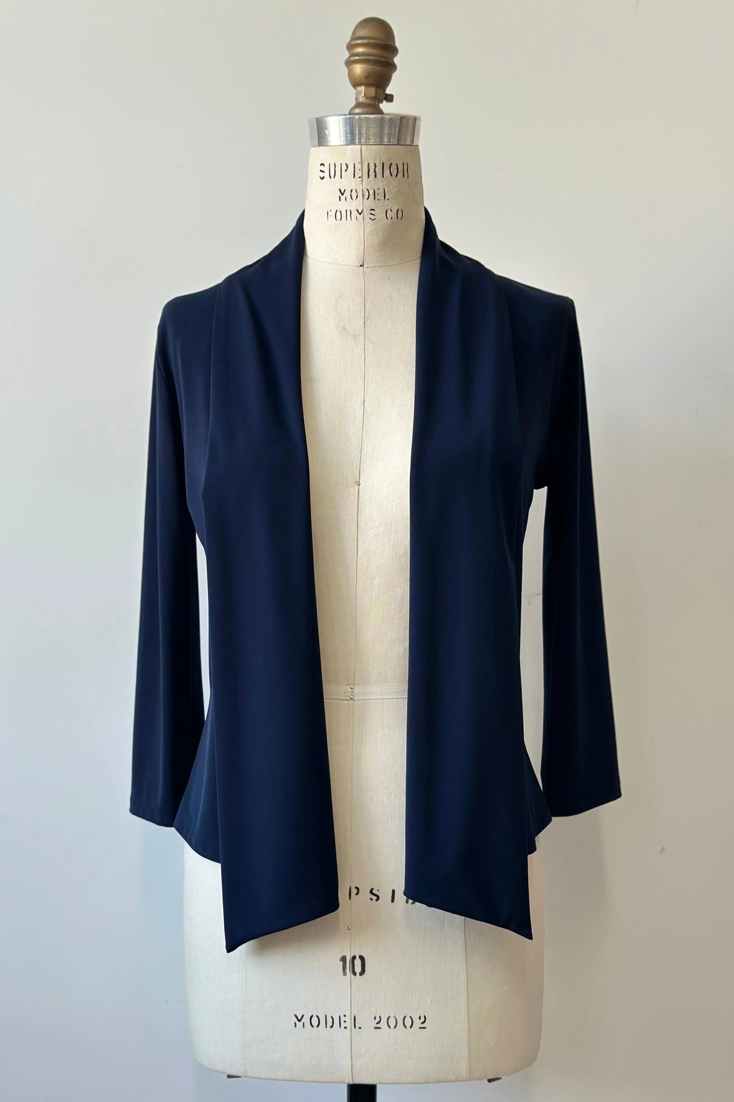 The Lioren Cardigan by Kollontai in Navy, a stretchy cropped jacket with longer pointed panels in the front that can be worn loose or knotted, is shown on a mannequin in front of a white background. 