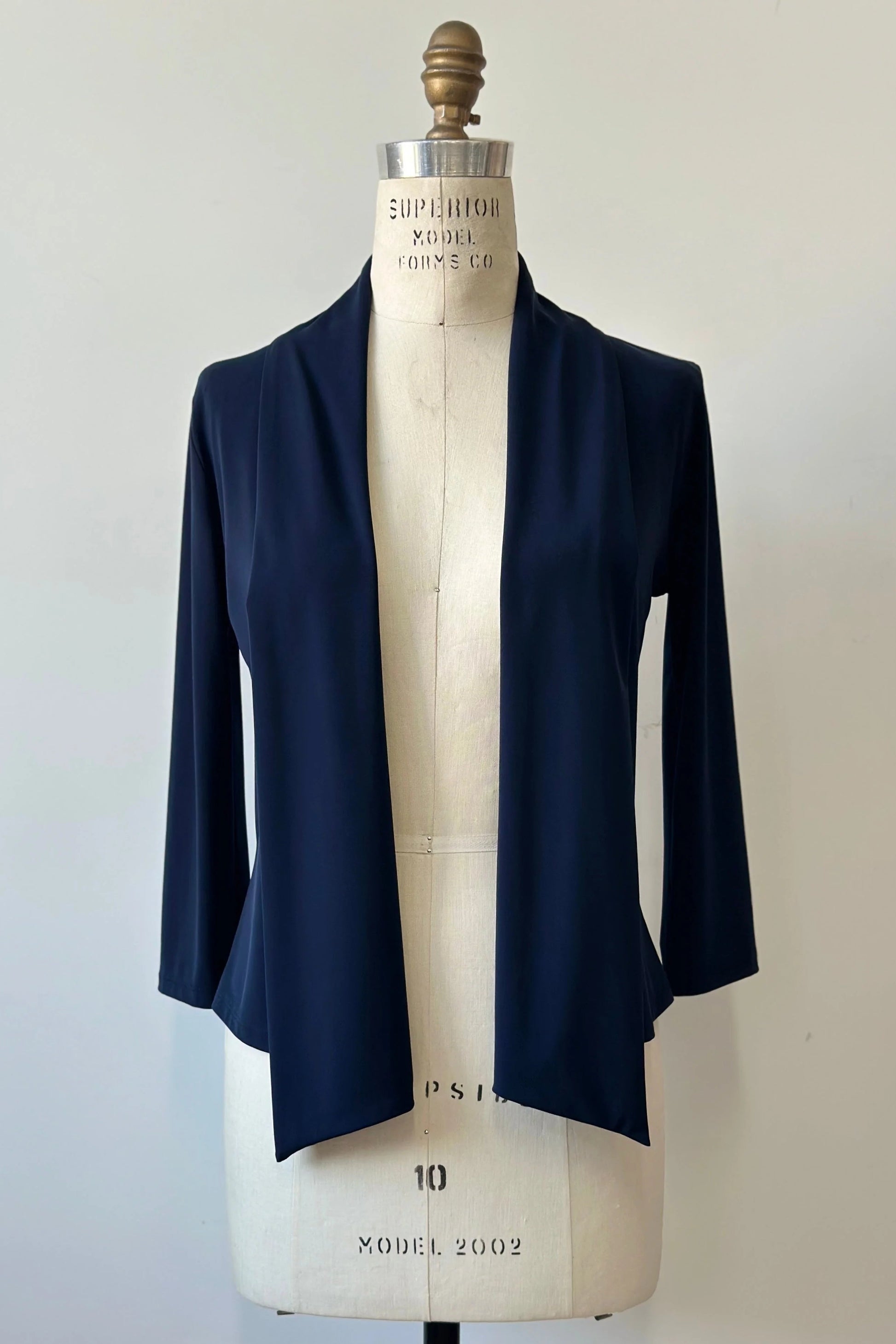 The Lioren Cardigan by Kollontai in Navy, a stretchy cropped jacket with longer pointed panels in the front that can be worn loose or knotted, is shown on a mannequin in front of a white background. 