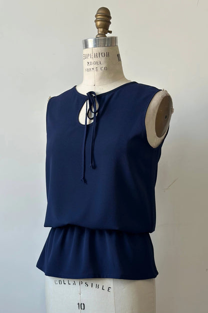 The Serena Top from Kollontai in Navy, a sleeveless top with a keyhole and tie neckline, elastic waist and flared peplum hem, is shown on a mannequin against a white background. 