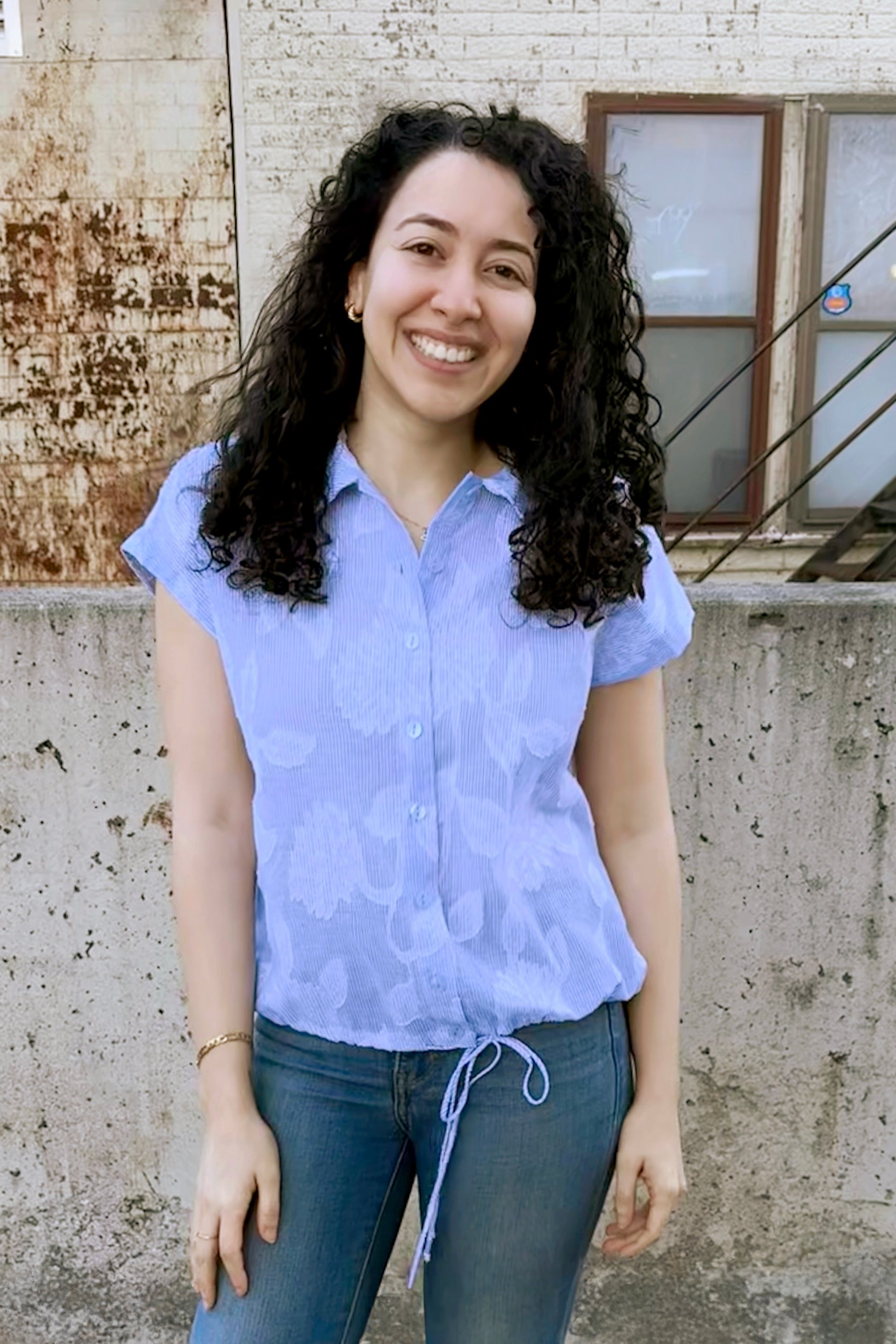 A woman wearing the Griffin Top by Pure Essence in Blue/White print, with a textured stripe/lace print, a short-sleeved blouse with a classic collar, button front, and drawstring detail at the waist.She is wearing it with jeans and standing outside by a brick wall. 