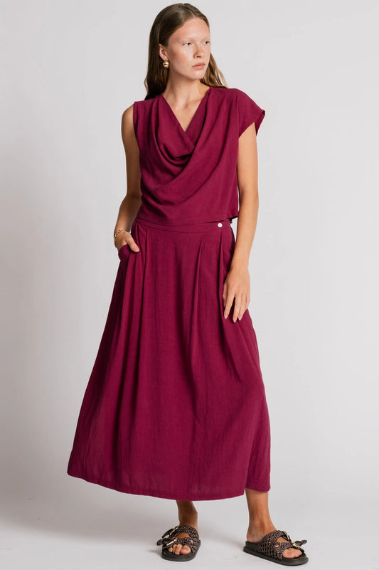 A woman wearing the Barbet Skirt by Allison Wonderland in Pink, a maxi dress with side slits, front pleats, elastic at the back waist and a side button detail at the front. She is wearing it with a matching tank and standing in front of a white background. 