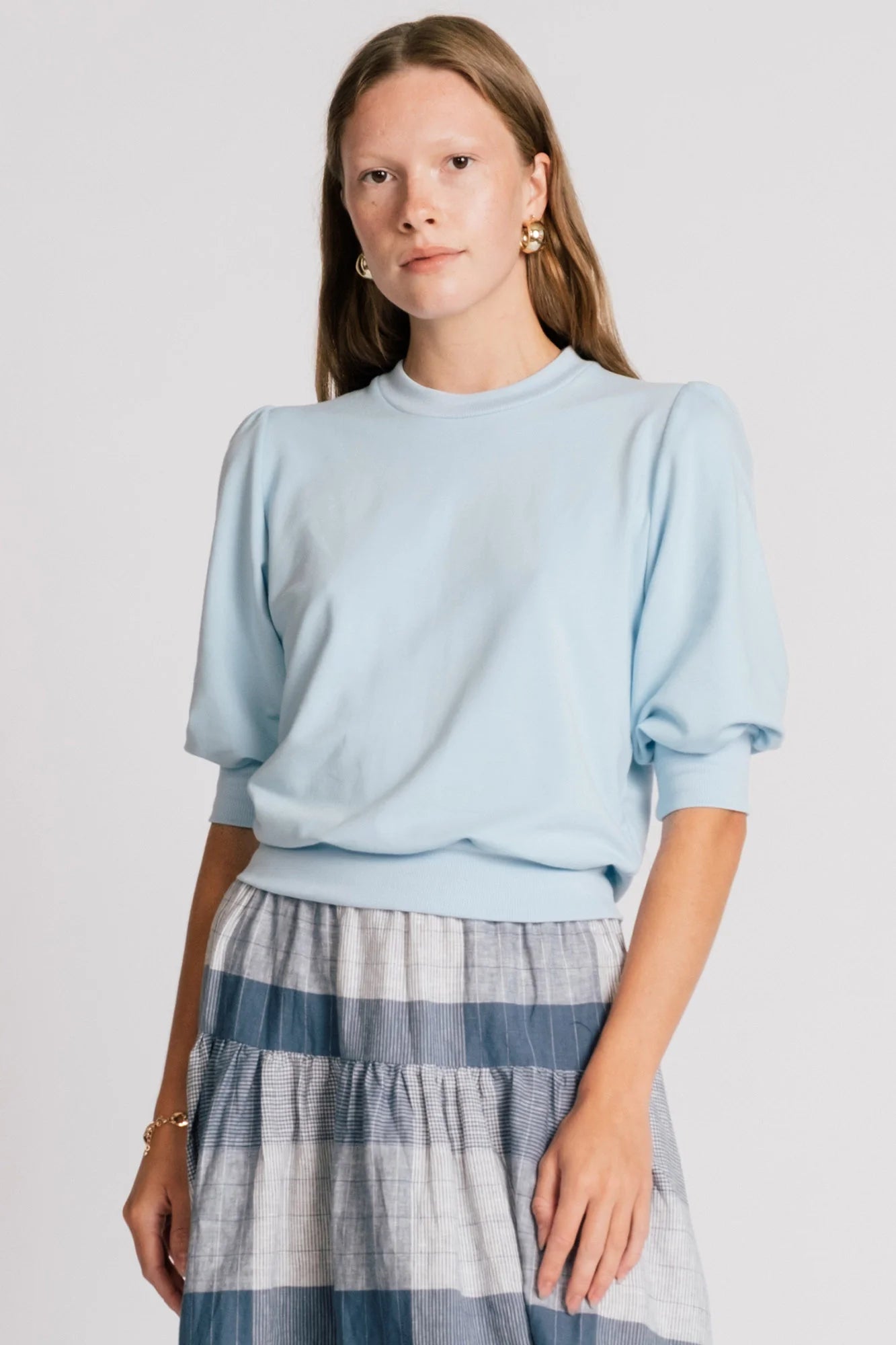 A woman wearing the Finch Sweatshirt by Allison Wonderland in Baby Blue, a cropped sweatshirt with elbow-length puffed sleeves and ribbing at the neck and waistband. She is wearing it with a blue plaid skirt and standing in front of a white background.