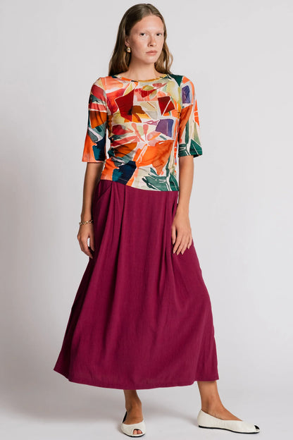 A woman wearing the Lorikeet Top by Allison Wonderland in multicolour print, with a high neck in front, a scooped neck at the back, and 3/4 sleeves. She is wearing it with a burgundy skirt and standing in front of a plain background