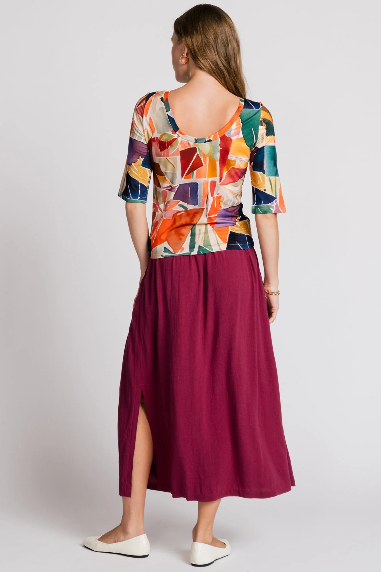 Back view of a woman wearing the Lorikeet Top by Allison Wonderland in multicolour print, with a high neck in front, a scooped neck at the back, and 3/4 sleeves. She is wearing it with a burgundy skirt and standing in front of a plain background