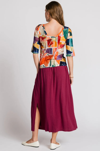 Back view of a woman wearing the Lorikeet Top by Allison Wonderland in multicolour print, with a high neck in front, a scooped neck at the back, and 3/4 sleeves. She is wearing it with a burgundy skirt and standing in front of a plain background