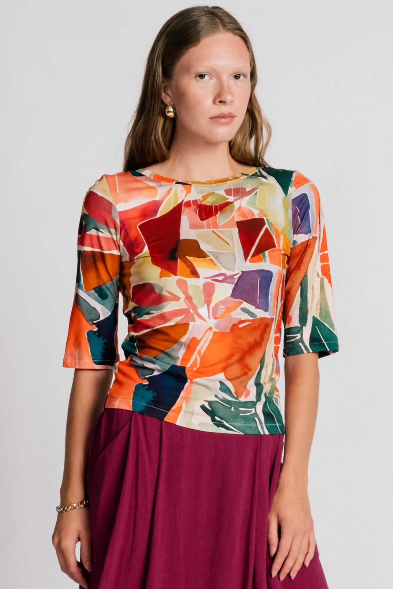 A woman wearing the Lorikeet Top by Allison Wonderland in multicolour print, with a high neck in front, a scooped neck at the back, and 3/4 sleeves. She is wearing it with a burgundy skirt and standing in front of a plain background