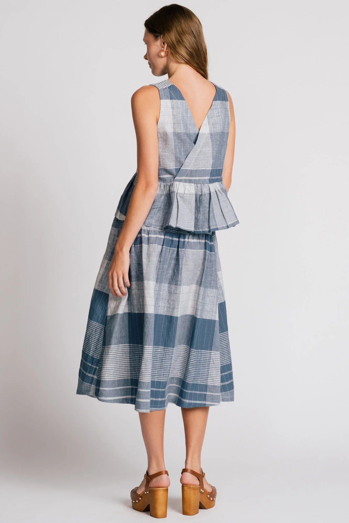 Back view of a woman wearing the Osprey Blouse by Allison Wonderland in Blue Plaid, a sleeveless top with a peplum both front and back and a cross-over back. She is wearing it with the matching Pelican Skirt and standing in front of a plain background. 