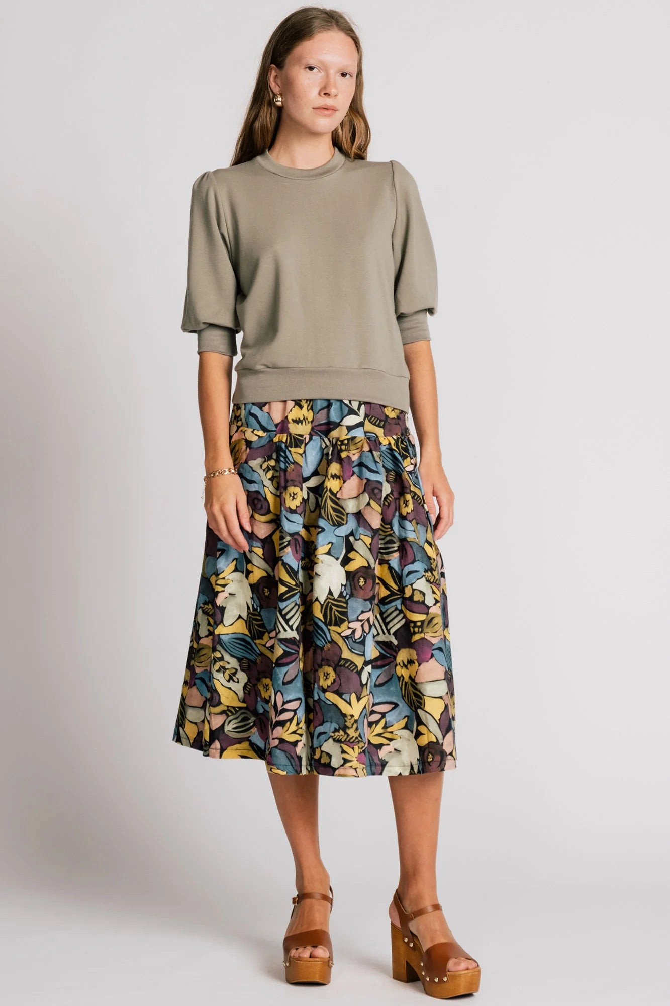 A woman wearing the Pelican skirt by Allison Wonderland in Black Floral, a mid-calf length tiered skirt with an elastic waist. She is wearing it with the Finch Sweatshirt in Eucalyptus and standing in front of a plain background. 
