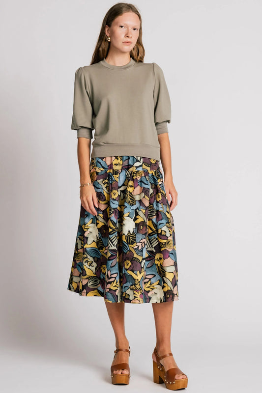 A woman wearing the Pelican skirt by Allison Wonderland in Black Floral, a mid-calf length tiered skirt with an elastic waist. She is wearing it with the Finch Sweatshirt in Eucalyptus and standing in front of a plain background. 