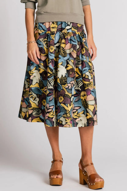 A waist down view of a woman wearing the Pelican skirt by Allison Wonderland in Black Floral, a mid-calf length tiered skirt with an elastic waist. She is wearing it with the Finch Sweatshirt in Eucalyptus and standing in front of a plain background. 