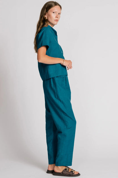 Side view of a woman wearing the Sparrow pants by Allison Wonderland in Ocean, straight-ish pants with an elastic waist. She is wearing them with the matching Kite Top  and standing in front of a plain background. 