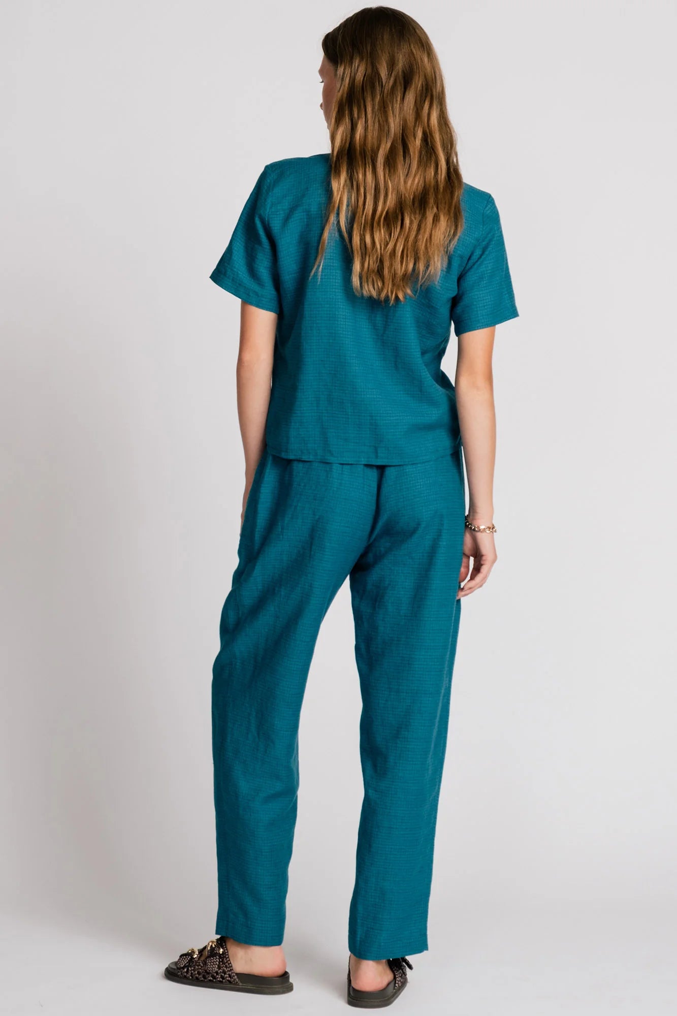Back view of a woman wearing the Sparrow pants by Allison Wonderland in Ocean, straight-ish pants with an elastic waist. She is wearing them with the matching Kite Top  and standing in front of a plain background. 