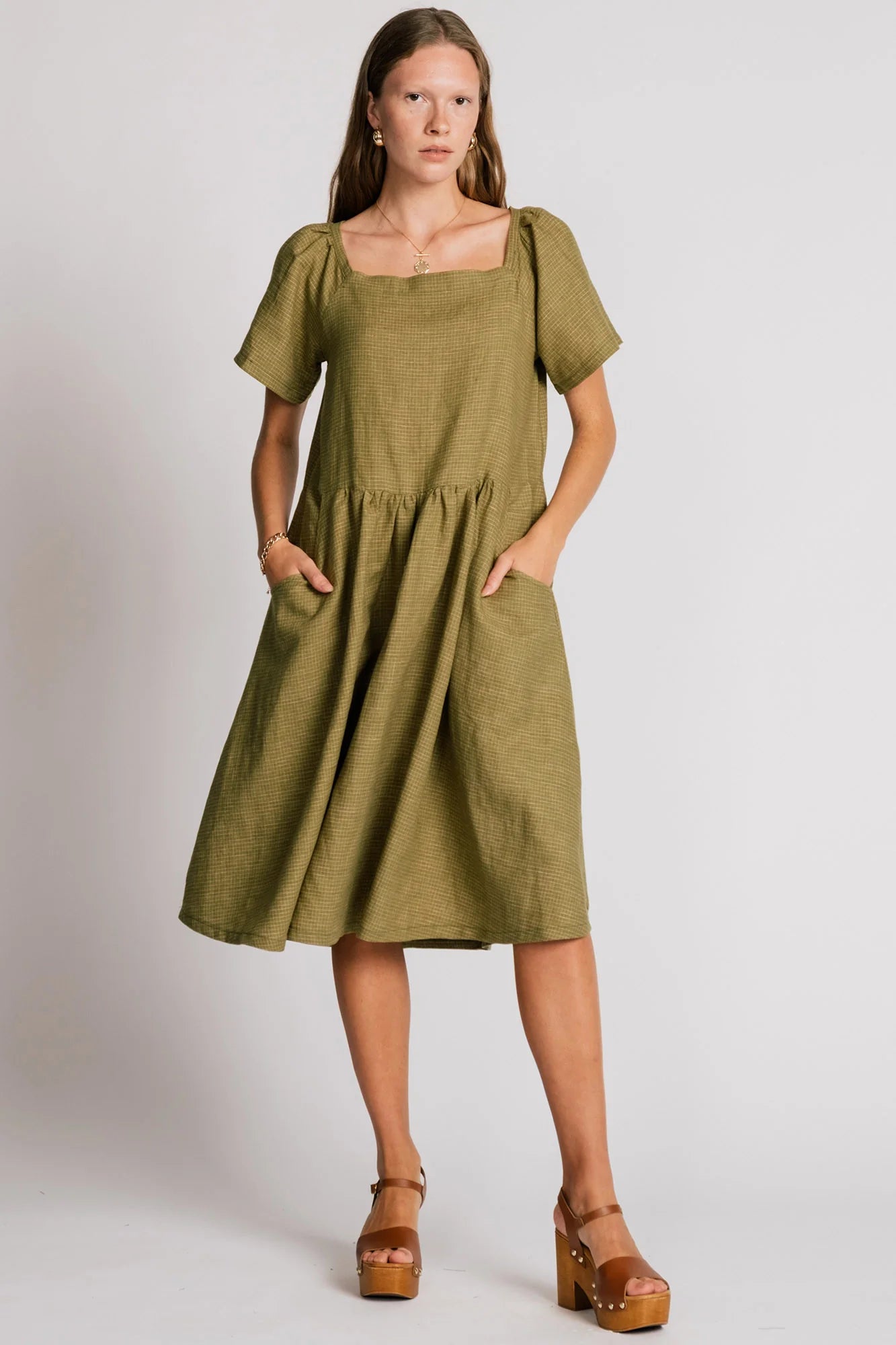 Woman wearing the Warbler Dress by Allison Wonderland in Avocado, a mid-calf length dress with short puffed sleeves and a square neckline. She is standing in front of a plain background. 
