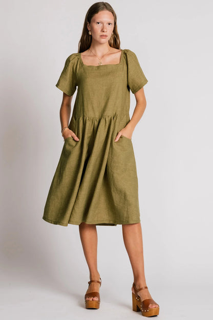 Woman wearing the Warbler Dress by Allison Wonderland in Avocado, a mid-calf length dress with short puffed sleeves and a square neckline. She is standing in front of a plain background. 
