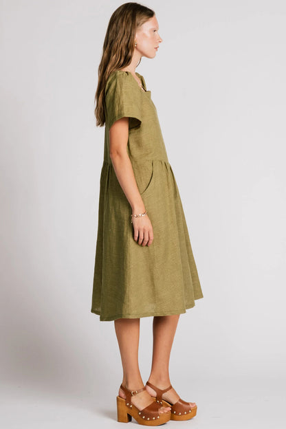 Side view of a woman wearing the Warbler Dress by Allison Wonderland in Avocado, a mid-calf length dress with short puffed sleeves and a square neckline. She is standing in front of a plain background. 