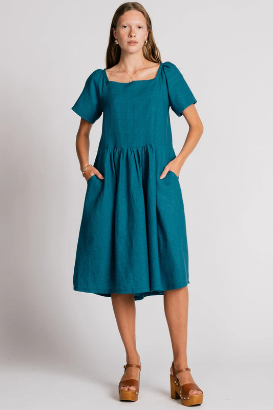 Woman wearing the Warbler Dress by Allison Wonderland in Ocean, a mid-calf length dress with short puffed sleeves and a square neckline. She is standing in front of a plain background. 
