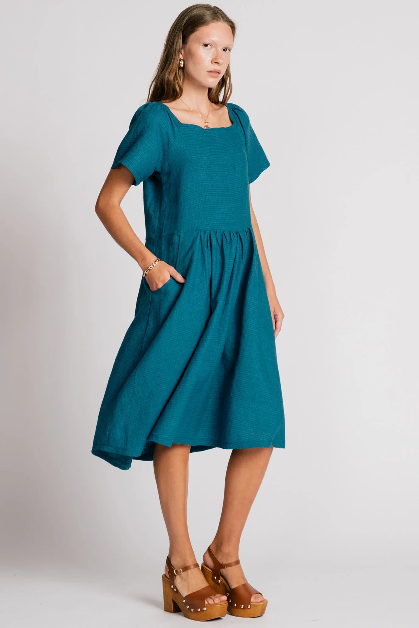 Woman wearing the Warbler Dress by Allison Wonderland in Ocean, a mid-calf length dress with short puffed sleeves and a square neckline. She is standing in front of a plain background. 