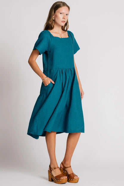 Woman wearing the Warbler Dress by Allison Wonderland in Ocean, a mid-calf length dress with short puffed sleeves and a square neckline. She is standing in front of a plain background. 