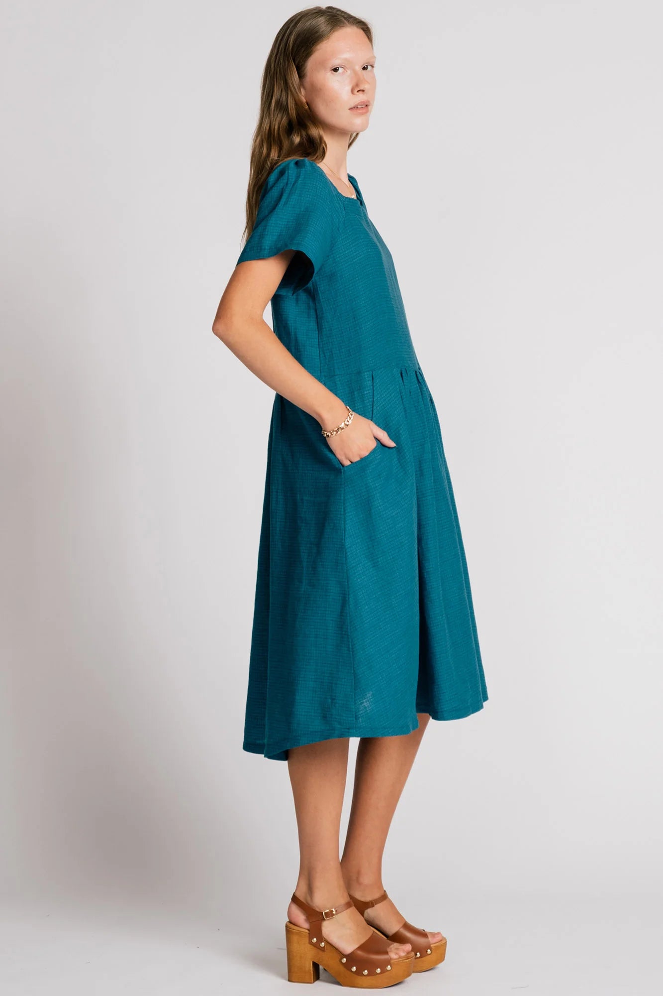Side view of woman wearing the Warbler Dress by Allison Wonderland in Ocean, a mid-calf length dress with short puffed sleeves and a square neckline. She is standing in front of a plain background. 