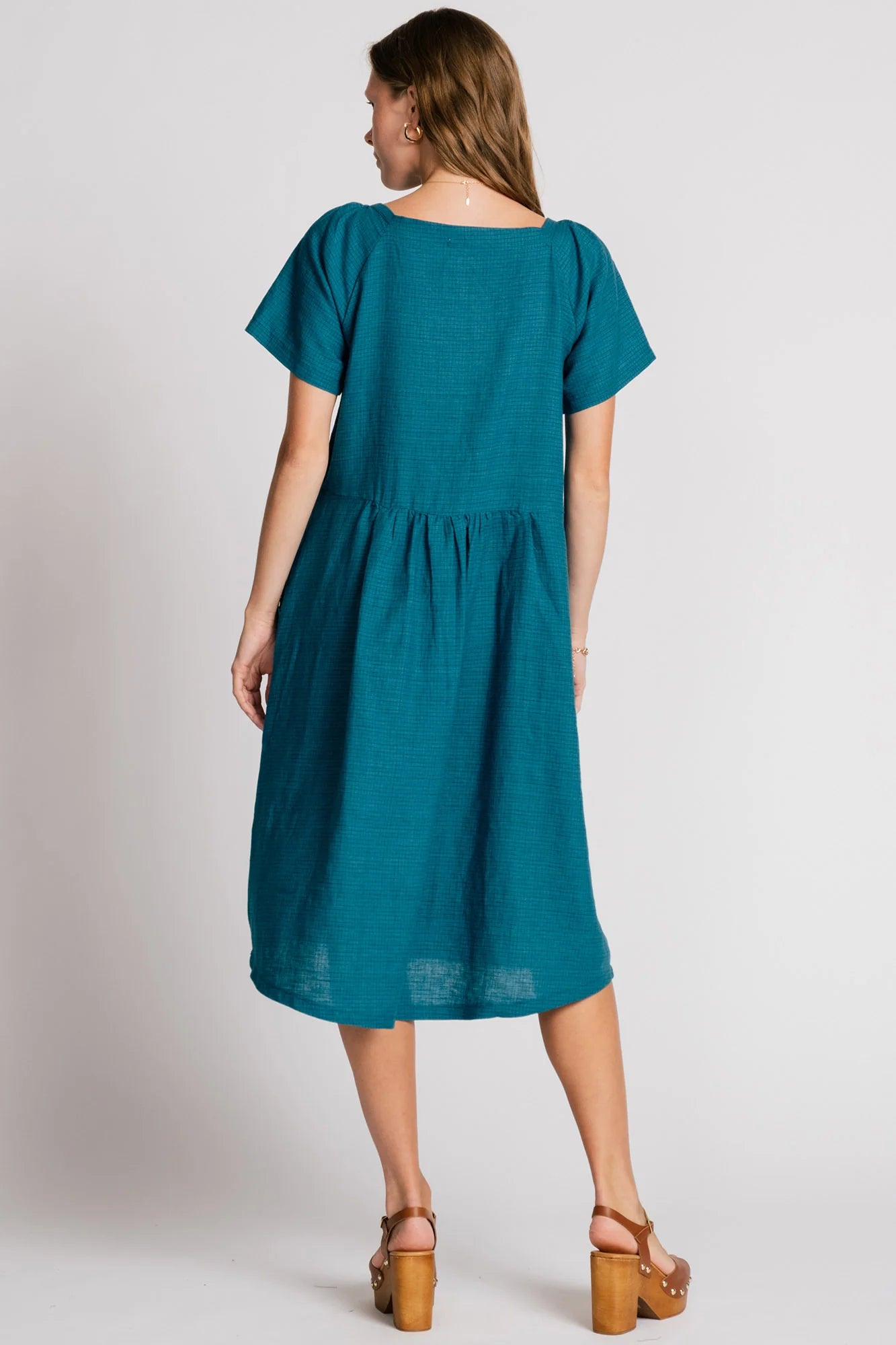 Back view of a woman wearing the Warbler Dress by Allison Wonderland in Ocean, a mid-calf length dress with short puffed sleeves and a square neckline. She is standing in front of a plain background. 