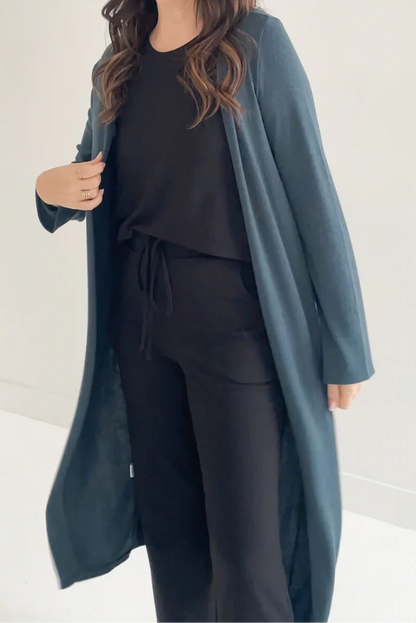 Neck to knee view of a woman wearing the Carolina Duster Cardigan by Ang Hill in Juniper, a long open cardigan with long sleeves. She is wearing it with black pants and a black top and is standing in front of a white background. 