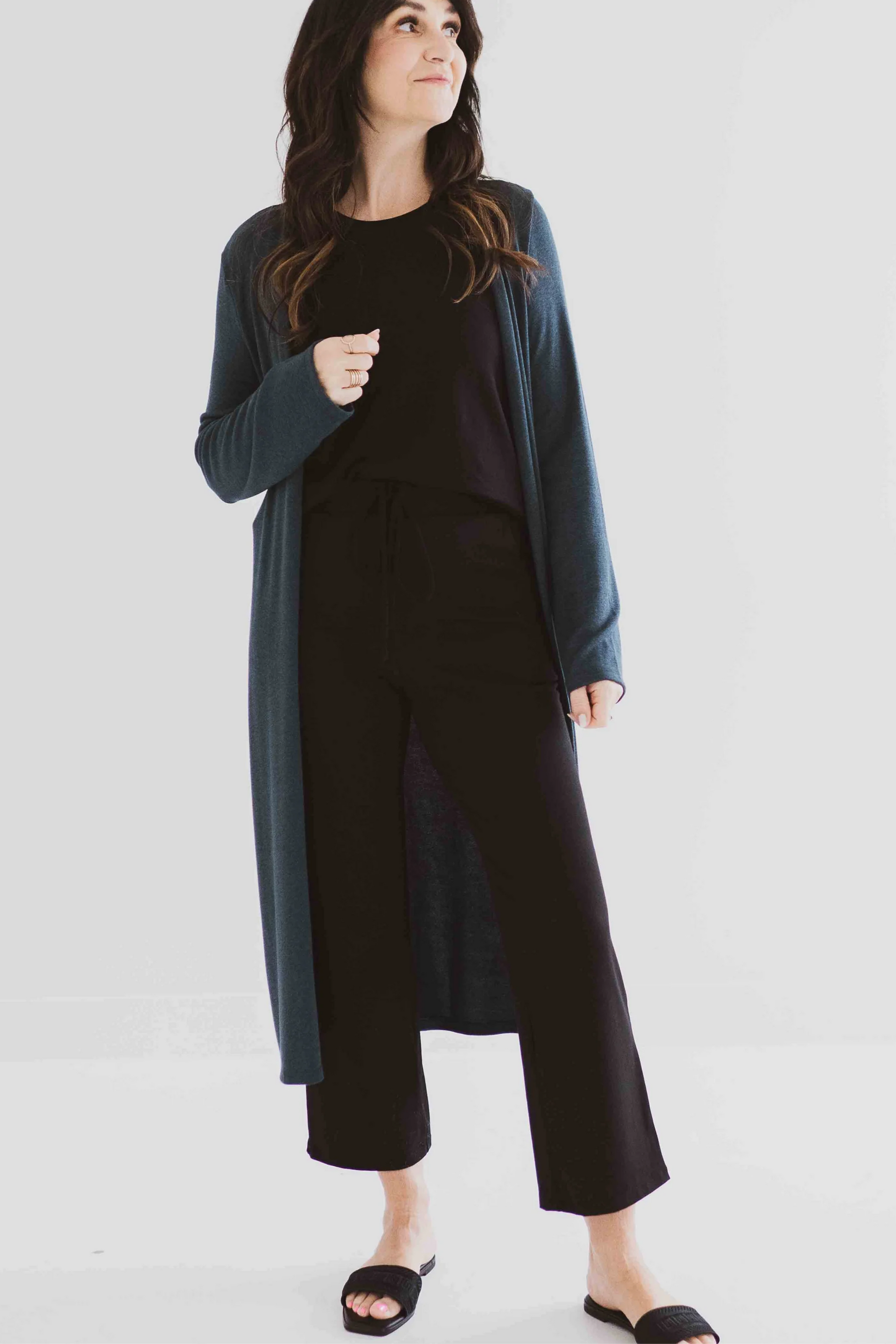 A woman wearing the Carolina Duster Cardigan by Ang Hill in Juniper, a long open cardigan with long sleeves. She is wearing it with black pants and a black top and is standing in front of a white background. 