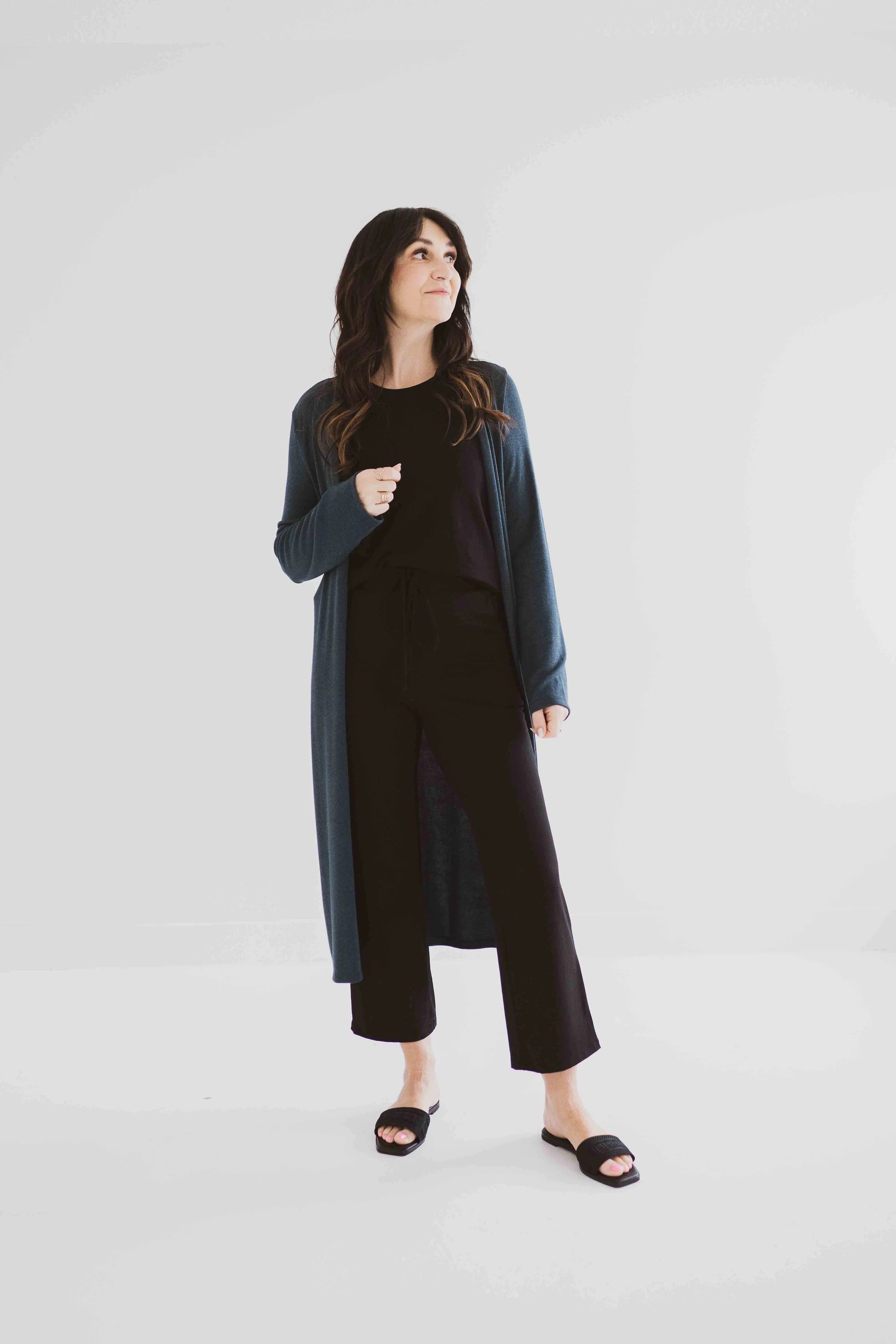 A woman wearing the Carolina Duster Cardigan by Ang Hill in Juniper, a long open cardigan with long sleeves. She is wearing it with black pants and a black top and is standing in front of a white background. 