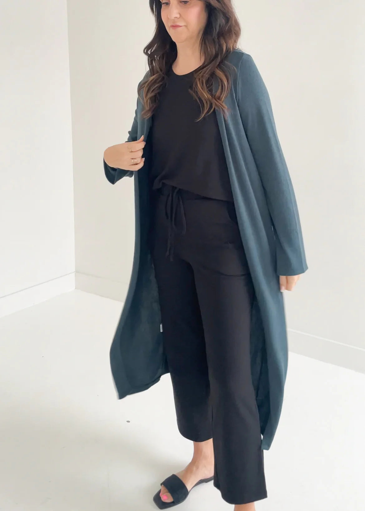 A woman wearing the Carolina Duster Cardigan by Ang Hill in Juniper, a long open cardigan with long sleeves. She is wearing it with black pants and a black top and is standing in front of a white background. 