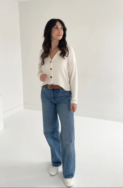 A woman wearing the Holly Cropped Cardigan by Ang Hill in Vanilla, a v-neck cardigan with drop sleeves and tortoiseshell buttons. She is wearing it with jeans and is standing in front of a white background. 