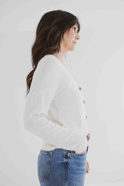 Side 3/4 view of a woman wearing the Holly Cropped Cardigan by Ang Hill in Vanilla, a v-neck cardigan with drop sleeves and tortoiseshell buttons. She is wearing it with jeans and is standing in front of a white background. 
