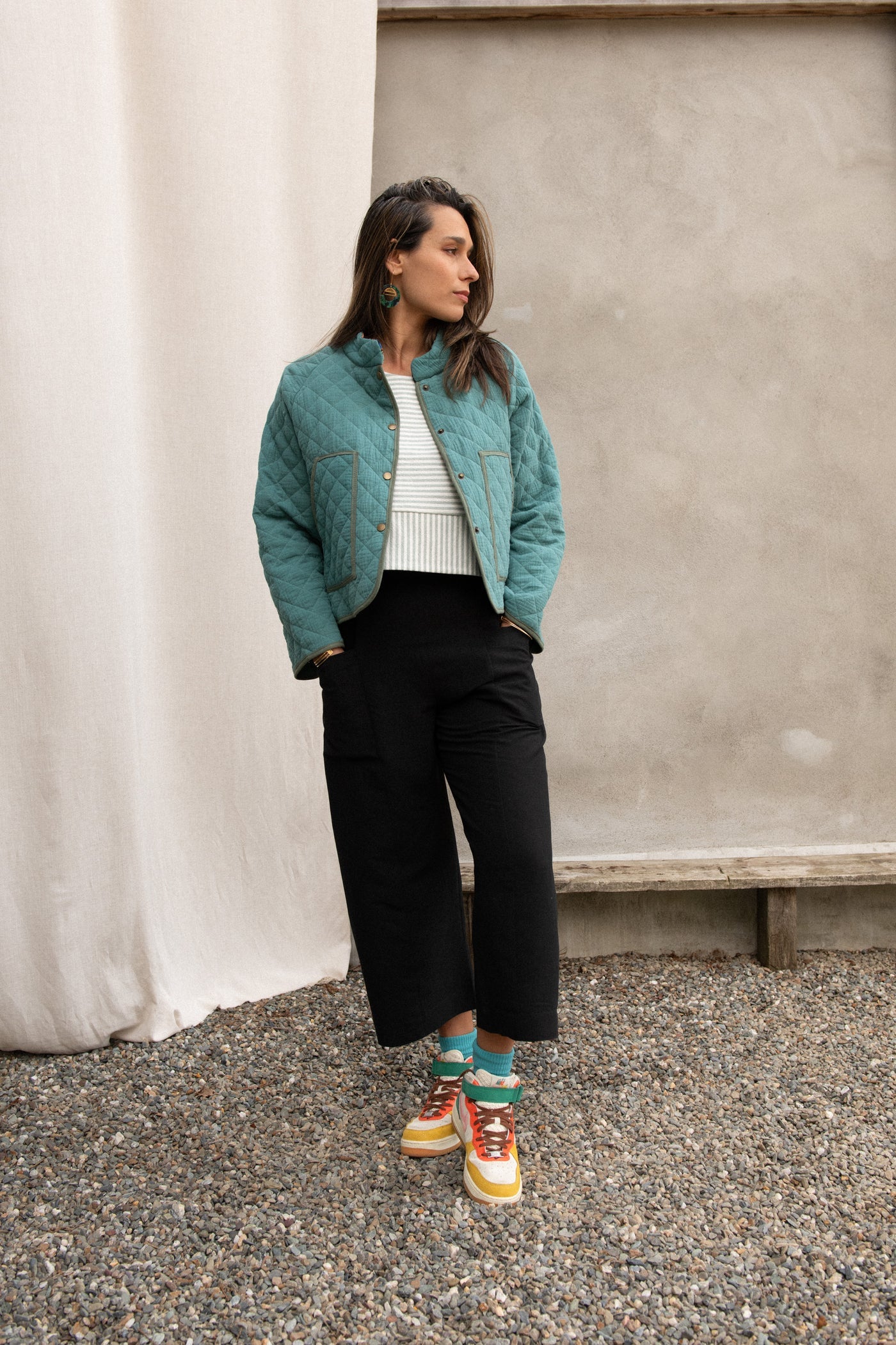 A woman wearing the Artemesia Jacket by Kazak in Mint, a quilted jacket with snaps up the front and large patch pockets. She is wearing it with a striped top and black pants and standing outside in front of a grey wall.