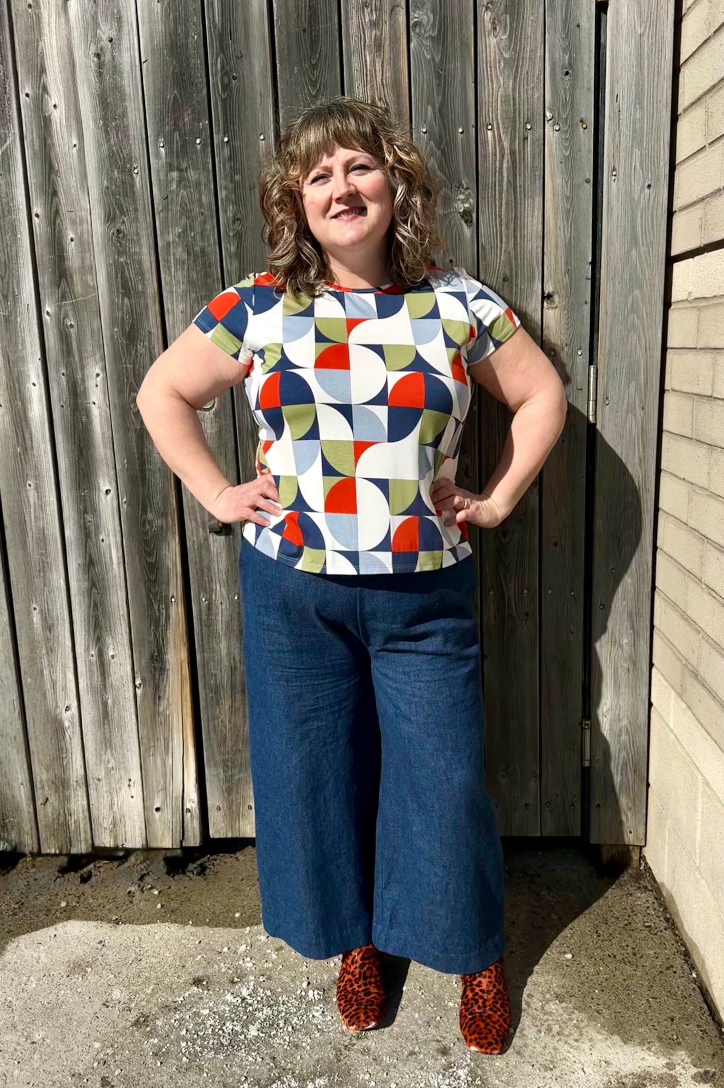 A woman wearing the Gwen Top by Pure Essence in Multicolour Mosaic with the Mateo pants by Melow,  standing against a wooden fence.
