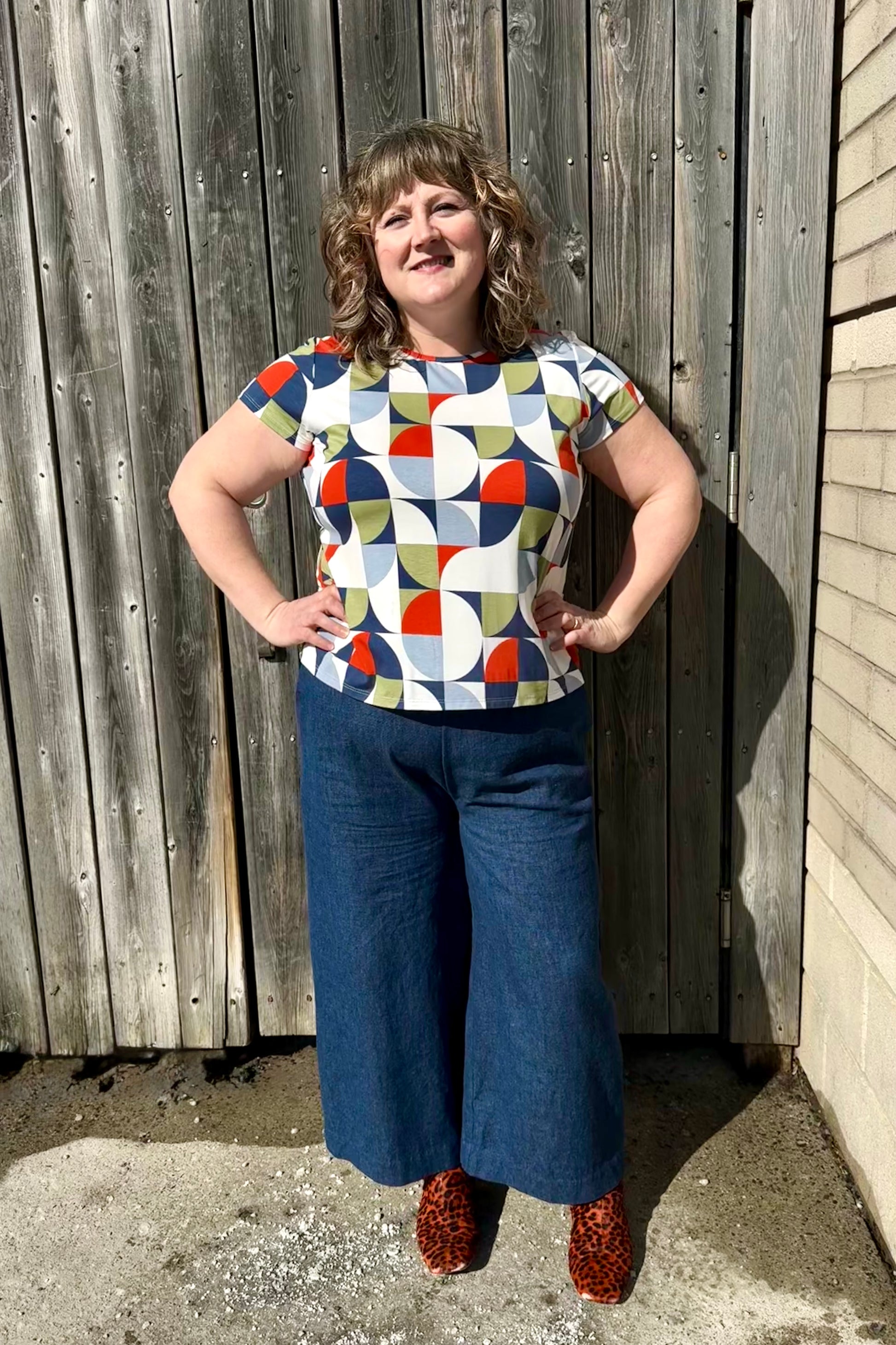A woman wearing the Gwen Top by Pure Essence in Multicolour Mosaic with the Mateo pants by Melow,  standing against a wooden fence.