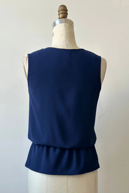 A back view of the Serena Top from Kollontai in Navy, a sleeveless top with a keyhole and tie neckline, elastic waist and flared peplum hem, is shown on a mannequin against a white background. 