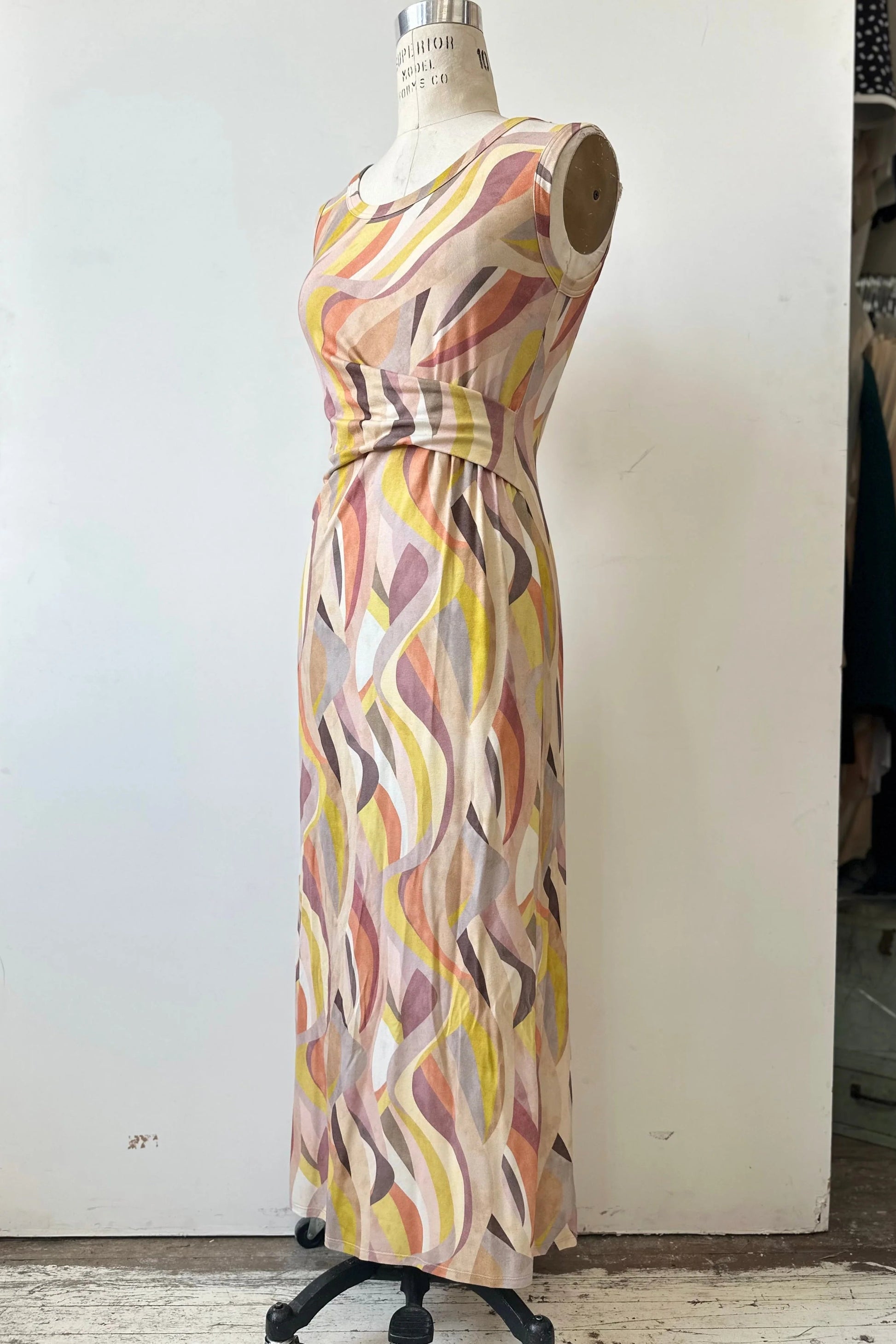 The Serina Dress by Kollontai in Peach print, a long, slim cut sleeveless dress with a soft pleat at the waist, is shown on a mannequin in front of a white background. 