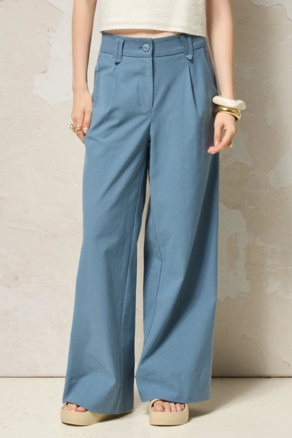 Waist down view of a woman wearing  the Chain pants from Cokluch in Ocean, with a slightly low waist, pointed belt loops, front pleats, and wide legs. She is wearing them with a white top  and standing against a grey background. 