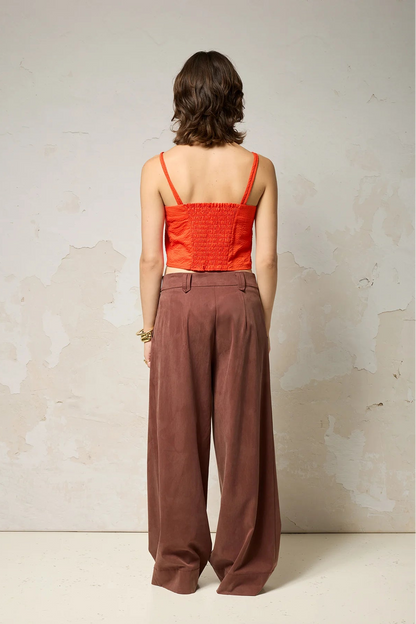 Back view of a woman wearing  the Chain pants from Cokluch in Cappuccino, with a slightly low waist, pointed belt loops, front pleats, and wide legs. She is wearing them with an orange top  and standing against a grey background. 