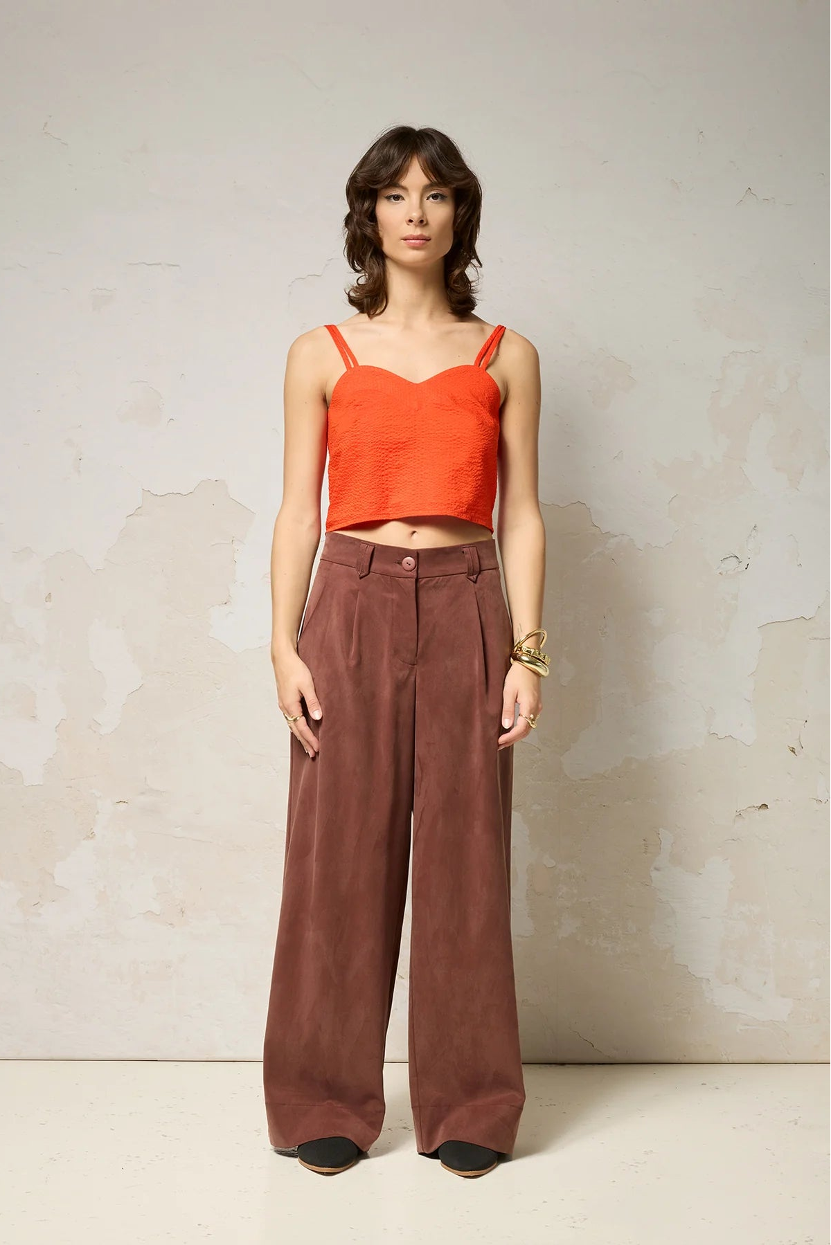 A woman wearing  the Chain pants from Cokluch in Cappuccino, with a slightly low waist, pointed belt loops, front pleats, and wide legs. She is wearing them with an orange top  and standing against a grey background. 