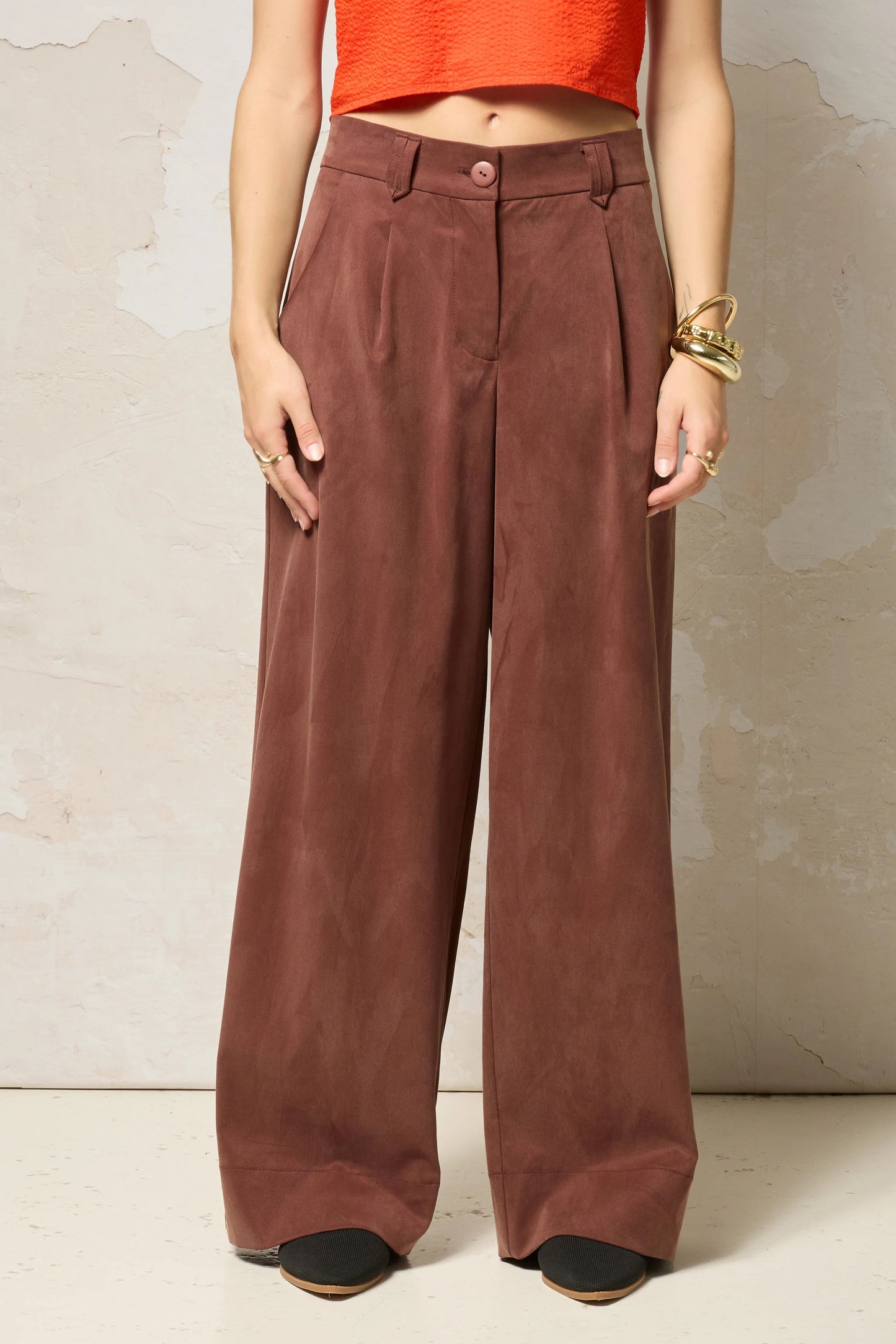 Waist down view of a woman wearing  the Chain pants from Cokluch in Cappuccino, with a slightly low waist, pointed belt loops, front pleats, and wide legs. She is wearing them with an orange top  and standing against a grey background. 
